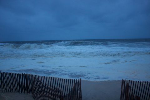 1990 Ford Bronco - A beach scene with a wooden fence in the foreground and large waves crashing on the shore under a stormy sky.