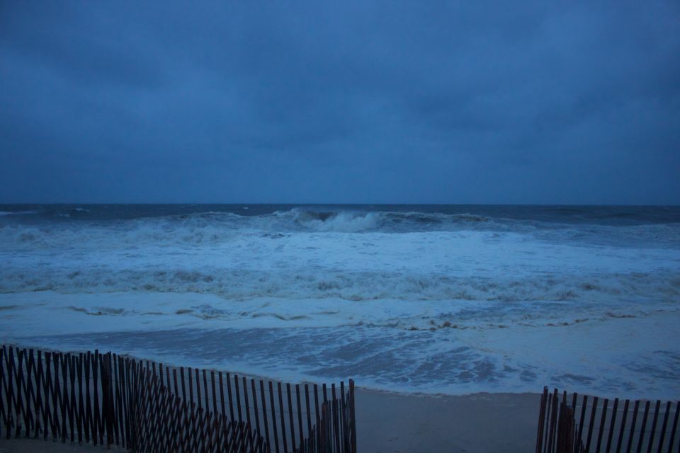 Stormy ocean waves crash on a beach behind a wooden fence. Supermotors.net logo visible