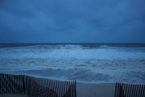 1990 Ford Bronco - Rough waves crash ashore under a dark, cloudy sky, with a fence lining the beach.