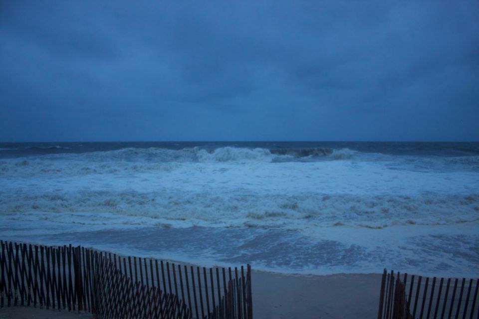 Stormy ocean waves crashing on a beach with a wooden fence in the foreground. Supermotors.net logo visible