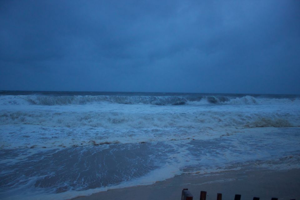Stormy ocean waves crash on a beach under a dark, cloudy sky. Watermark reads SUPERMOTORS