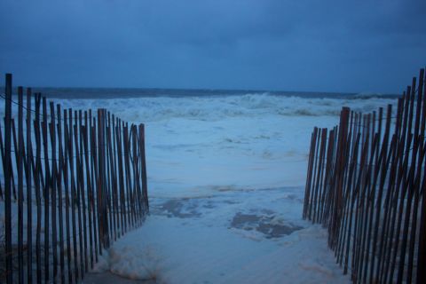 1990 Ford Bronco - A beach scene with rough ocean waves and foam, framed by a weathered wooden fence, under a dark, cloudy sky.