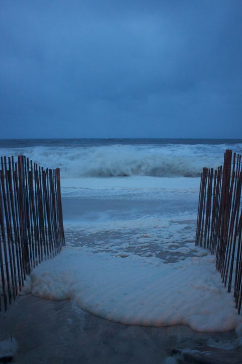 1990 Ford Bronco - A beach path is covered in sea foam with large waves crashing in the ocean under a dark, cloudy sky.