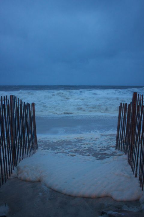 1990 Ford Bronco - Ocean waves crash onto a foamy beach, framed by wooden fence posts under a gloomy sky.
