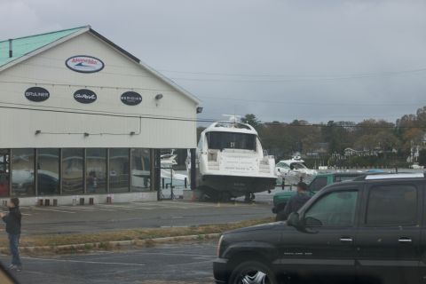 1990 Ford Bronco - A large white yacht is parked on a trailer outside a marina building, with several cars visible in the foreground.
