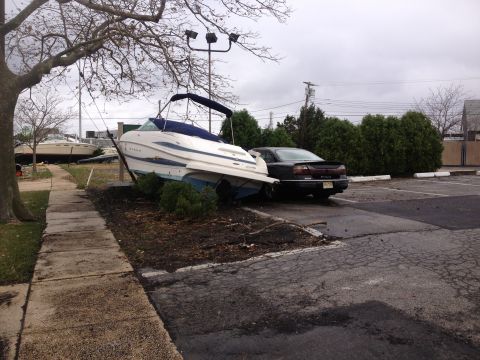 1990 Ford Bronco - Storm damage shows a boat displaced onto land next to a car in a parking lot.