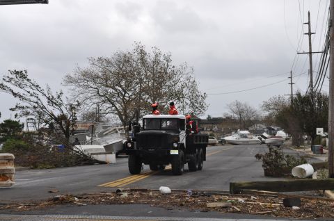 1990 Ford Bronco - A military truck navigates a road littered with debris and damaged boats, suggesting a post-disaster scene.