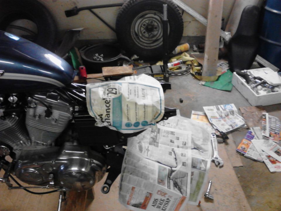 motorcycle engine and blue tank in a cluttered garage with parts and newspapers