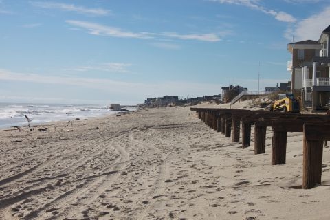 1990 Ford Bronco - A yellow excavator is visible on the beach near damaged pilings and houses, suggesting post-storm cleanup.