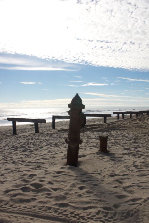 1990 Ford Bronco - A rusty fire hydrant stands partially buried in the sand on a beach, with wooden structures and the ocean behind it.