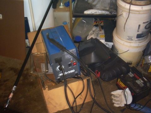 1991 Ford LTD Crown Victoria - A blue MIG-100 flux-core wire welder sits on a cardboard box in a cluttered garage, with its torch lead coiled on the floor beside a black…