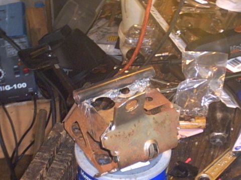 1991 Ford LTD Crown Victoria - A rusty cast-iron engine or suspension mounting bracket clamped in a vise on a workbench, with a MIG-100 wire-feed welder and cluttered sho…