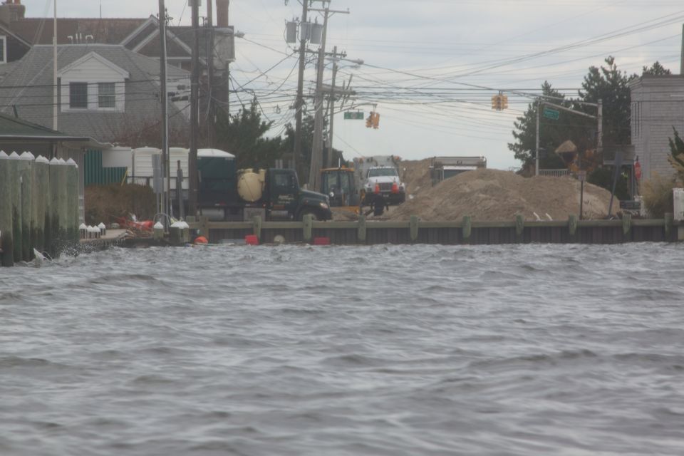 Several trucks and construction vehicles are parked on a pier with a pile of sand behind them, with choppy water in the foreground