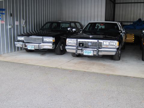 1991 Ford LTD Crown Victoria - Two black sedans, one a 1991 Ford Crown Victoria LTD, are parked side-by-side in a garage.
