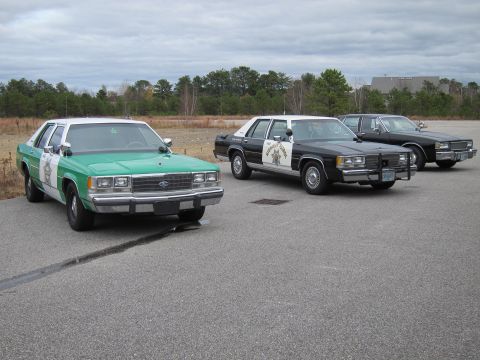 1991 Ford LTD Crown Victoria - Three police cars are parked on an asphalt lot under a cloudy sky.