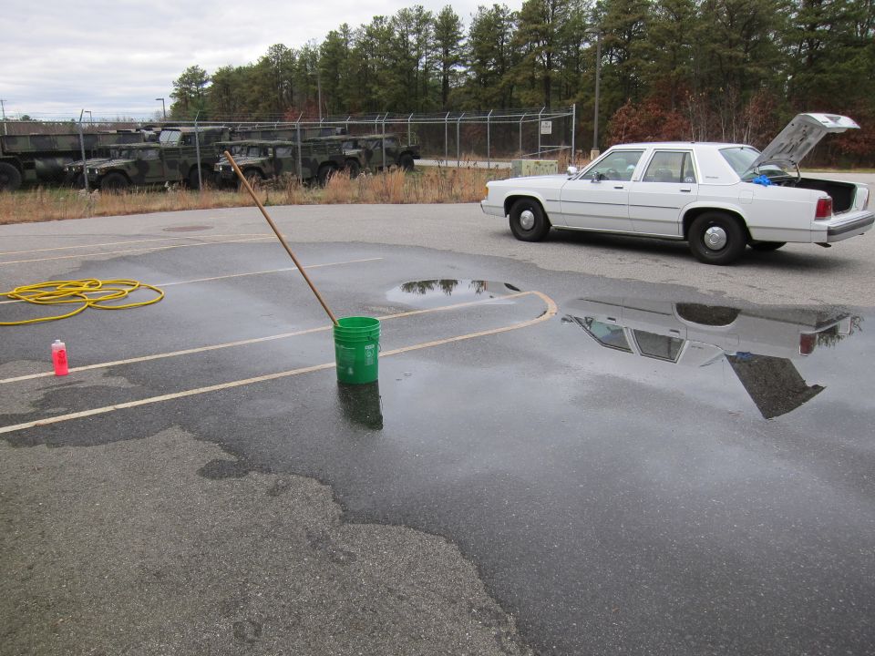 White sedan parked next to puddles, with military vehicles in background