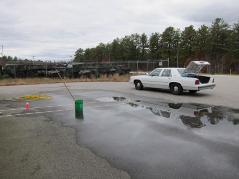 1991 Ford LTD Crown Victoria - A white sedan is parked in a lot with puddles, while military vehicles are visible behind a fence.