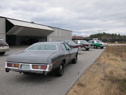 1991 Ford LTD Crown Victoria - Several older sedans are parked in a line next to a large garage building on a cloudy day.