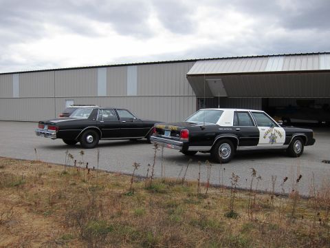 1991 Ford LTD Crown Victoria - Two police cruisers, one a 1991 Ford Crown Victoria LTD, are parked outside a large building with an open bay door.