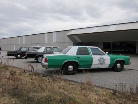 1991 Ford LTD Crown Victoria - Three older model police sedans are parked in a row outside a large building with an open hangar door.