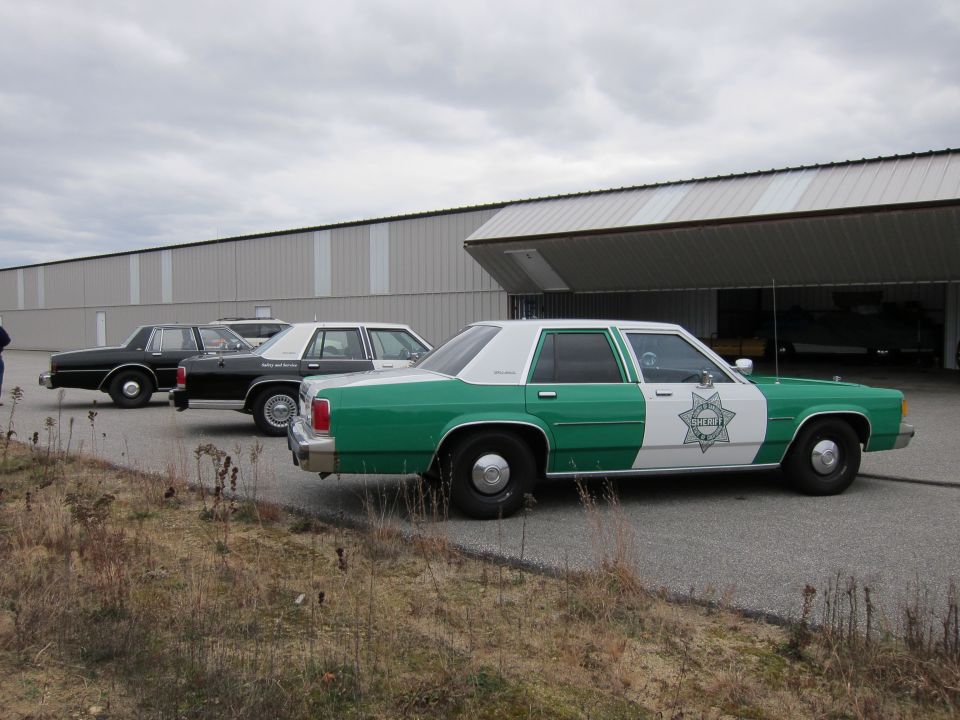 Three police cars, one green and white, two black, parked on asphalt in front of a large building with an open bay