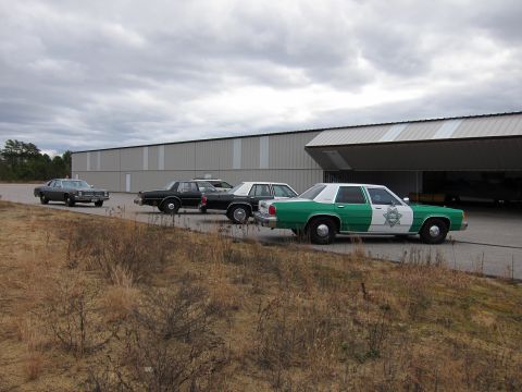1991 Ford LTD Crown Victoria - Several sedans, including a green and white police car, are parked in front of a large building with an open bay door.