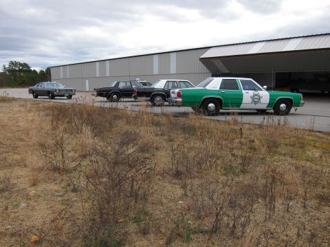 1991 Ford LTD Crown Victoria - Four police cars are parked in a line next to a large building with an open bay door.