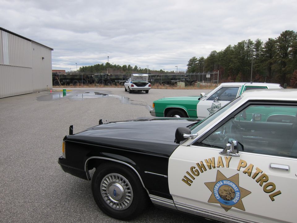 Black and white highway patrol car in foreground, green and white patrol car behind it, white car with open trunk in distance