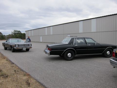 1991 Ford LTD Crown Victoria - Two sedans are parked on asphalt in front of a large industrial building.