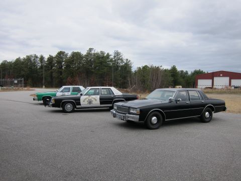 1991 Ford LTD Crown Victoria - Three older sedans are parked in a lot. One appears to be a police car with visible text on its door.
