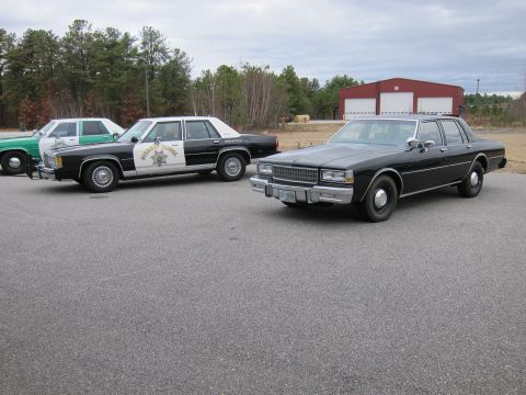 1991 Ford LTD Crown Victoria - A black sedan is parked in front of a police car and a green car, with a red building in the background.
