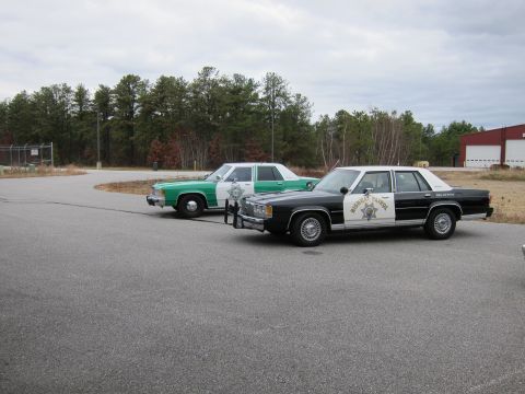 1991 Ford LTD Crown Victoria - Two police cars, one black and white and one green and white, are parked in an asphalt lot.
