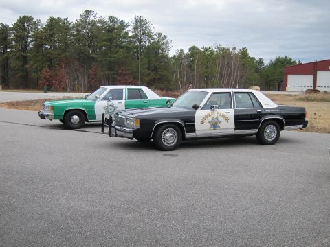 1991 Ford LTD Crown Victoria - Two older model police sedans are parked side-by-side in a lot with trees and a building in the background.