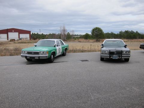 1991 Ford LTD Crown Victoria - Two police sedans, likely from the 1980s, are parked outdoors on a cloudy day.