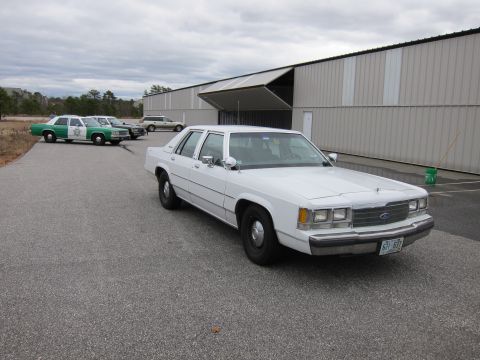 1991 Ford LTD Crown Victoria - A white 1991 Ford Crown Victoria LTD is parked in front of a large industrial building, with other vehicles visible in the background.