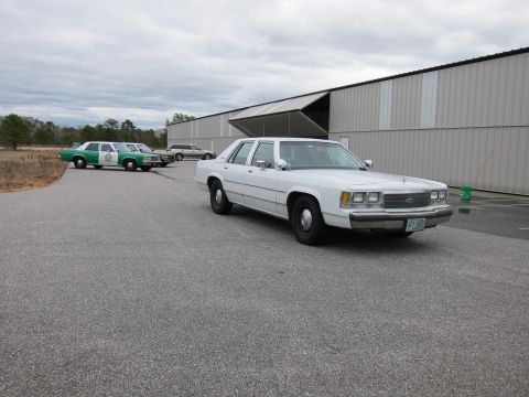 1991 Ford LTD Crown Victoria - A white sedan is parked in front of a large building with other vehicles in the background.
