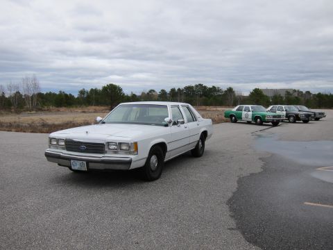 1991 Ford LTD Crown Victoria - A white 1991 Ford Crown Victoria LTD is parked in an open lot, with several other vehicles visible in the background.