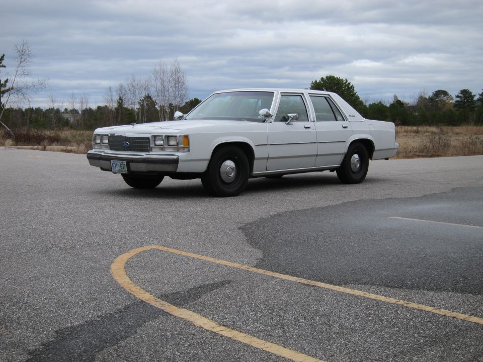 White Ford Crown Victoria parked on asphalt with yellow lines. Trees and cloudy sky in background