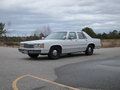 1991 Ford LTD Crown Victoria - A white sedan is parked on an asphalt lot under a cloudy sky. The vehicle's front end is visible from a 3/4 angle.