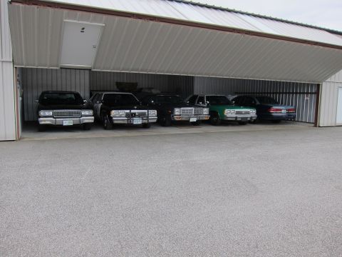 1991 Ford LTD Crown Victoria - Multiple vehicles are parked inside a large open garage with a metal roof and walls.