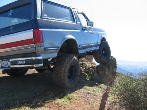 1989 Ford Bronco - This lifted Ford Bronco is shown off-road, with its front tires on rocks and a scenic view in the background.