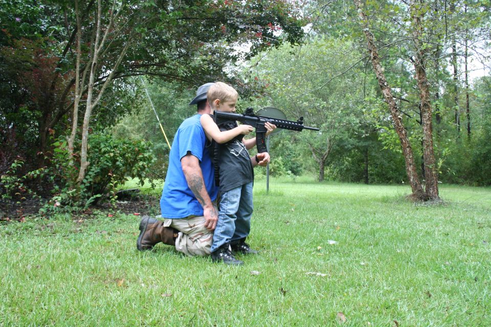 Man in blue shirt and tan pants kneels as a child in a black shirt holds a rifle