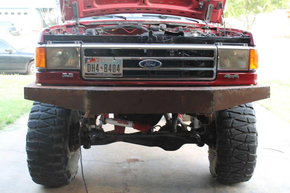 Red Ford truck front view with custom metal bumper, large tires, and open hood