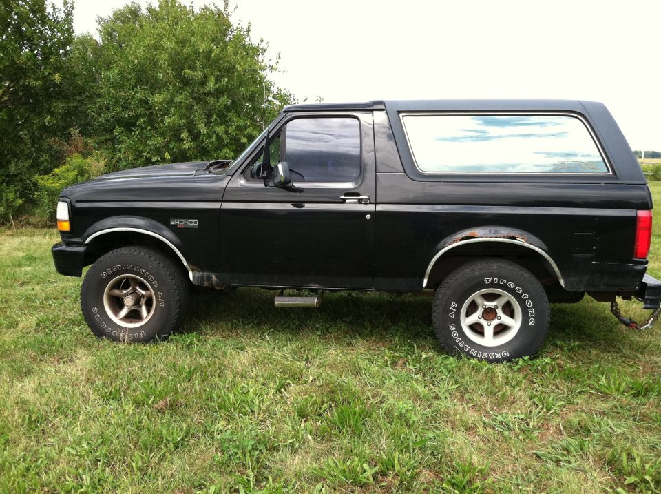 Side view of a black Ford Bronco parked on grass with trees in the background. The Bronco has chrome trim and aftermarket wheels
