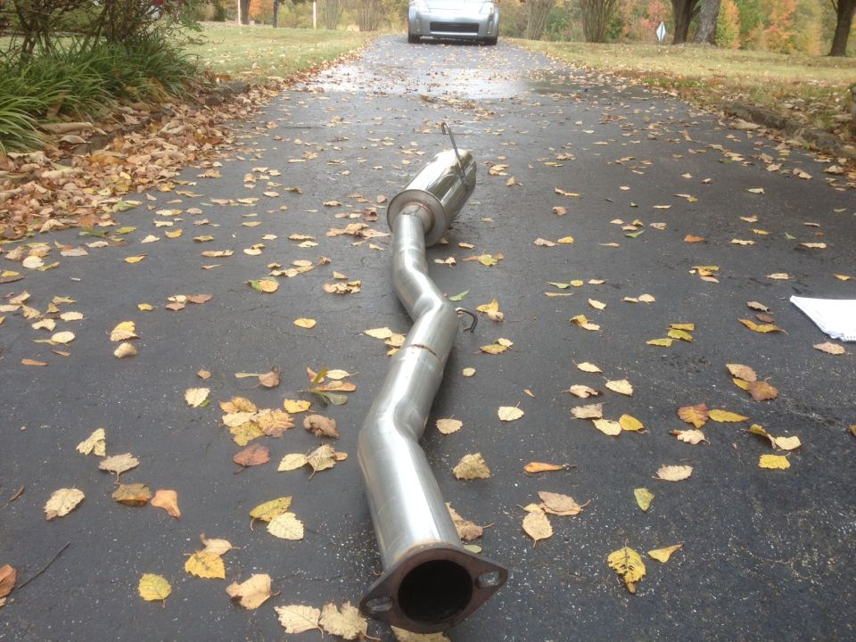 Exhaust pipe on asphalt driveway with fallen leaves. A silver car is visible in the distance