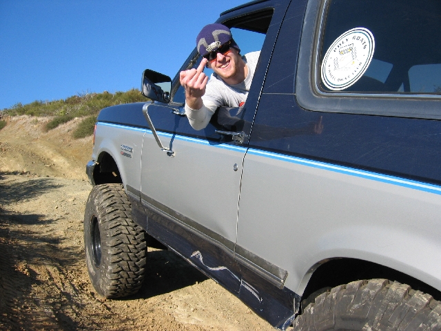Blue and silver lifted Ford Bronco on dirt trail with driver leaning out window