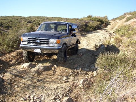 1989 Ford Bronco - A Ford Bronco navigates a challenging, sun-drenched off-road path with sparse vegetation.