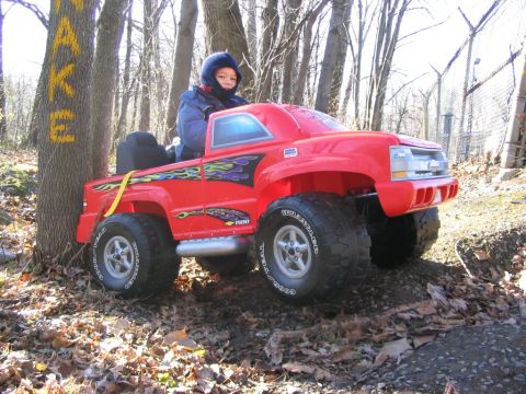 1986 Ford Bronco - A child sits in a red toy truck with flame decals, parked amongst trees and fallen leaves.