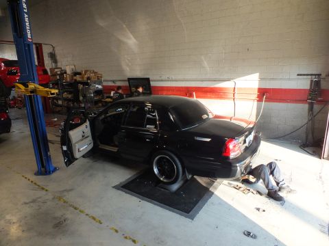 2003 Ford Crown Victoria - Black sedan running on a garage dyno next to a blue lift, a worker lying beneath the chassis
