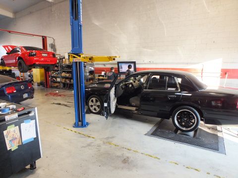 2003 Ford Crown Victoria - Black full-size sedan staged on a chassis dyno, driver door open, red car raised on a rear lift